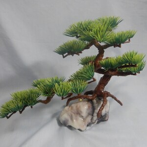 Op de afbeelding: Een miniatuur kunstmatige bonsai boom met groene bladeren en een bruine stam. De boom is gemonteerd op een lichtgekleurde, getextureerde rotsbasis. De takken strekken zich horizontaal uit, waardoor een evenwichtige en esthetisch aantrekkelijke compositie ontstaat.