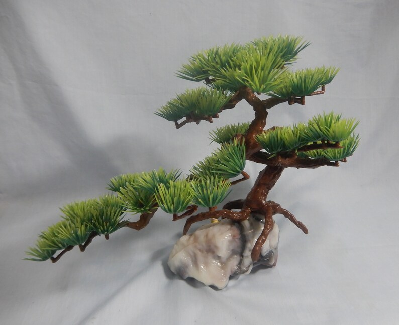 Op de afbeelding: Een miniatuur kunstmatige bonsai boom met groene bladeren en een bruine stam. De boom is gemonteerd op een lichtgekleurde, getextureerde rotsbasis. De takken strekken zich horizontaal uit, waardoor een evenwichtige en esthetisch aantrekkelijke compositie ontstaat.