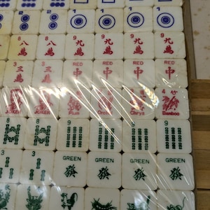 May include: A set of white Mahjong tiles with blue, red, and green markings. The tiles are arranged in rows and columns, some wrapped in plastic. The tiles feature various symbols, numbers, and Chinese characters. The set includes tiles marked "Joker", "Red", and "Green".
