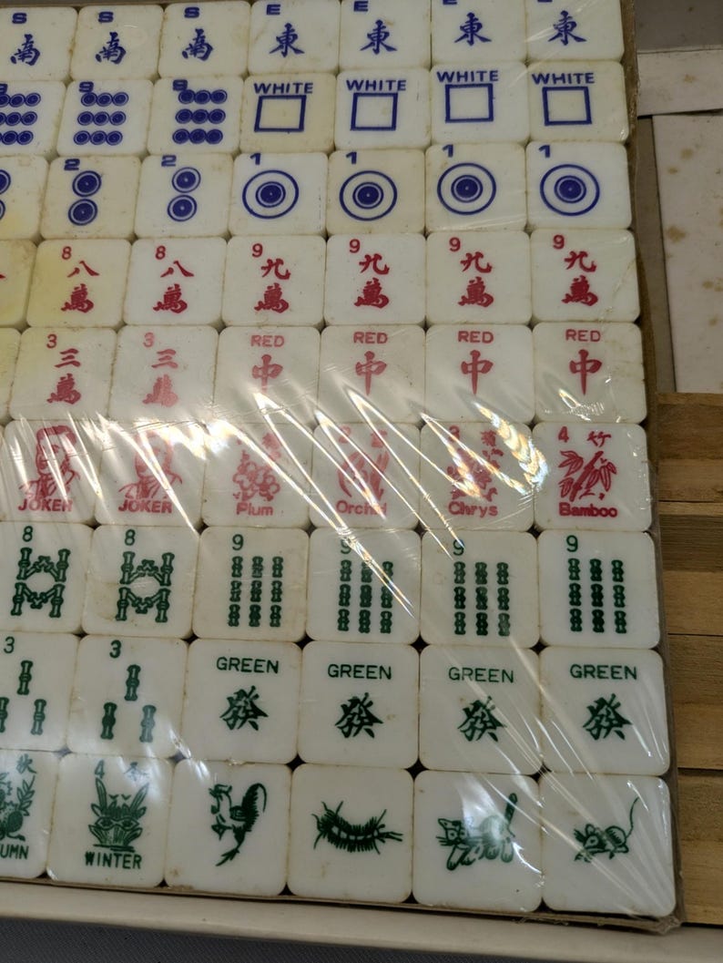 May include: A set of white Mahjong tiles with blue, red, and green markings. The tiles are arranged in rows and columns, some wrapped in plastic. The tiles feature various symbols, numbers, and Chinese characters. The set includes tiles marked "Joker", "Red", and "Green".