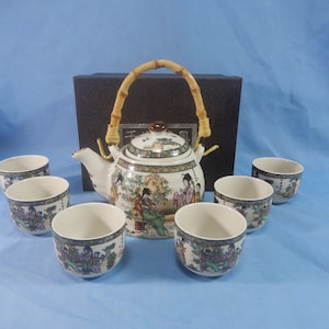Könnte beinhalten: Eine weiße Teekanne aus Keramik mit einem Bambusgriff und einem Blumendesign sowie fünf passende Teetassen. Die Teekanne und die Tassen haben ein farbenfrohes Blumendesign mit asiatisch inspirierten Figuren.