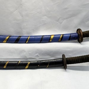 Könnte beinhalten: Zwei dekorative Katana-Schwerter. Eines ist blau mit goldenen und schwarzen Streifen, das andere ist schwarz mit goldenen Streifen. Beide haben braune Griffe, die mit dunklem Bindfaden umwickelt sind.