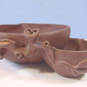 Puede incluir: Un macetero de cerámica de terracota con dos cuencos, uno más grande que el otro, conectados por un diseño de pájaro esculpido.
