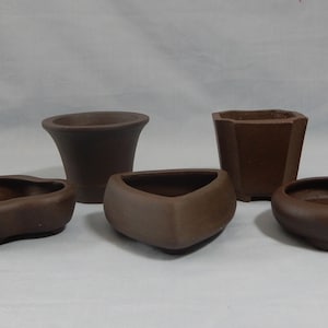May include: Five brown ceramic bonsai pots of different shapes and sizes. The pots are arranged in a row on a white surface.