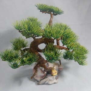 Op de afbeelding: Een miniatuur bonsai-boom met heldergroene bladeren en een getextureerde bruine stam. De boom staat op een decoratieve rotsbasis, met een klein beeldje van een persoon in gele gewaden. De algehele esthetiek doet denken aan traditionele Japanse kunst.