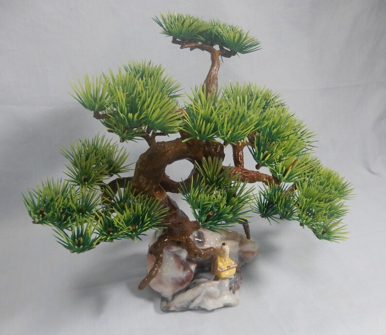 Op de afbeelding: Een miniatuur bonsai-boom met heldergroene bladeren en een getextureerde bruine stam. De boom staat op een decoratieve rotsbasis, met een klein beeldje van een persoon in gele gewaden. De algehele esthetiek doet denken aan traditionele Japanse kunst.