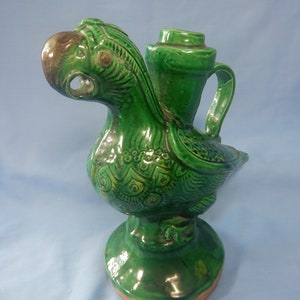 Puede incluir: Un recipiente verde de cerámica con forma de pájaro, con pico y asa. El pájaro tiene un diseño detallado y texturizado y un pico marrón. El recipiente está montado sobre una base redonda. El diseño general es ornamentado y decorativo.