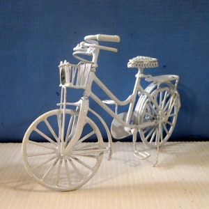 Könnte beinhalten: Ein weißes Metallfahrrad mit einem Korb und einem Sattel. Das Fahrrad hat einen weißen Rahmen und weiße Räder.