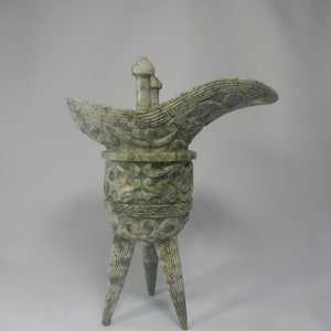 Puede incluir: Una copa ceremonial de jade verde con tres patas y un pico. La copa está decorada con intrincados tallados de dragones y otras criaturas míticas.