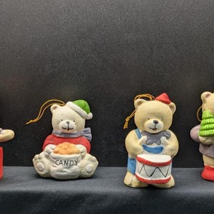 May include: Four ceramic bear ornaments with festive outfits. One bear is on a red toy box, another holds a candy bag, a third plays a drum, and the last holds a Christmas tree. Each has a gold string for hanging.
