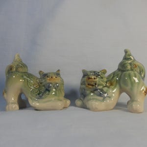 Może przedstawiać: Dwie ceramiczne figurki foo dog, jedna zielona i niebieska z uniesionym ogonem, druga zielona i niebieska z uniesioną głową. Obie mają białą glazurę i są skierowane do przodu.