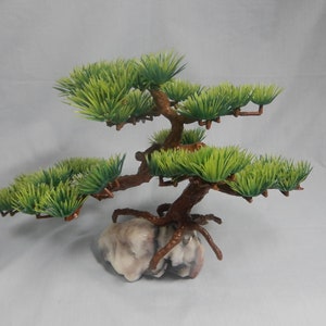 Op de afbeelding: Een kleine kunstmatige bonsai boom met groene bladeren en bruine takken. De boom staat op een lichtgekleurde, getextureerde rotsbasis, wat een natuurlijk uiterlijk geeft. Het ontwerp is bedoeld voor decoratieve doeleinden.