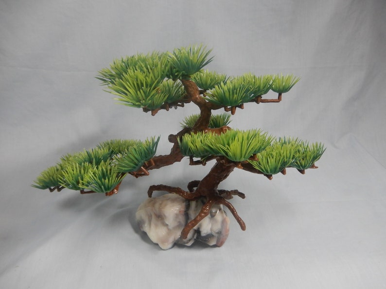 Op de afbeelding: Een kleine kunstmatige bonsai boom met groene bladeren en bruine takken. De boom staat op een lichtgekleurde, getextureerde rotsbasis, wat een natuurlijk uiterlijk geeft. Het ontwerp is bedoeld voor decoratieve doeleinden.