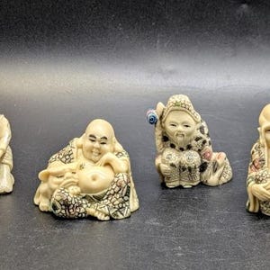 Pode incluir: Quatro pequenas estatuetas de cor branco-osso representando figuras sentadas em trajes tradicionais com padrões intrincados. As figuras apresentam expressões faciais detalhadas, incluindo uma com um cesto, uma com um pequeno animal, uma com um cajado e uma com as mãos juntas.