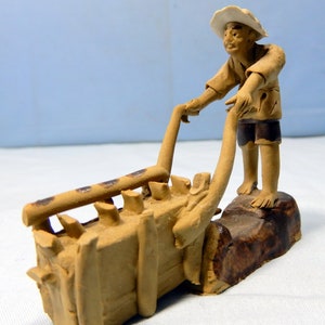 May include: A ceramic figurine of a person pushing a cart with a load of goods. The figurine is brown and has a white hat. The cart is made of wood and has a brown finish.