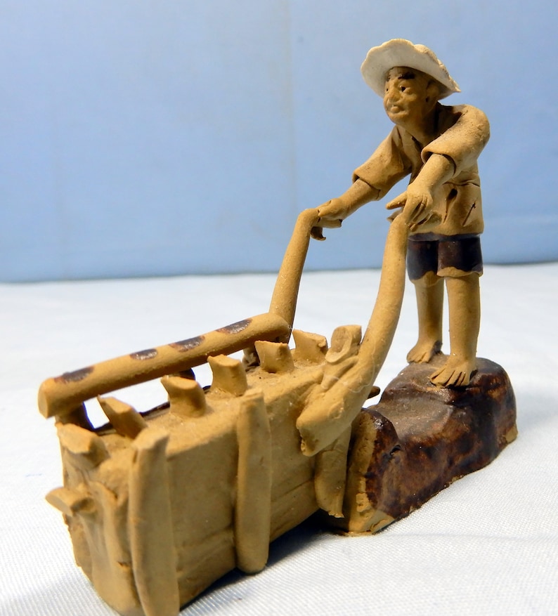 May include: A ceramic figurine of a person pushing a cart with a load of goods. The figurine is brown and has a white hat. The cart is made of wood and has a brown finish.