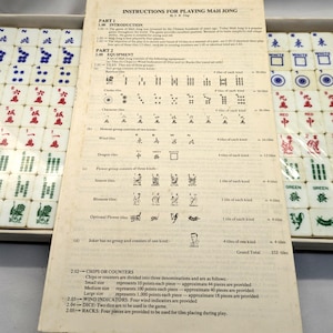 May include: A Mahjong game set with instructions. The set includes tiles with various symbols and characters, along with a booklet detailing the rules. The tiles are white with red, green, and blue markings. The game is in a box.