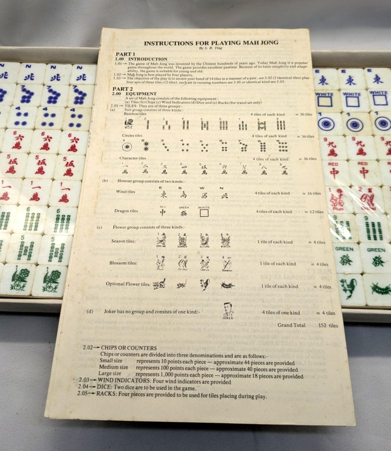 May include: A Mahjong game set with instructions. The set includes tiles with various symbols and characters, along with a booklet detailing the rules. The tiles are white with red, green, and blue markings. The game is in a box.