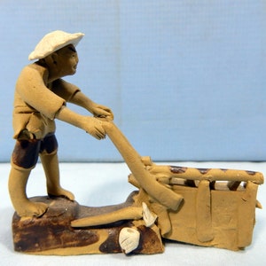 May include: A small ceramic figurine of a person in a straw hat pushing a wheelbarrow. The figurine is brown and has a rustic look.