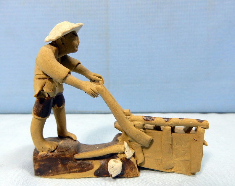 May include: A small ceramic figurine of a person in a straw hat pushing a wheelbarrow. The figurine is brown and has a rustic look.