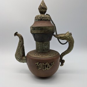 Puede incluir: Una tetera antigua de color cobre con detalles ornamentados de latón. La tetera presenta un asa y un pico en forma de dragón, un adorno decorativo y una cadena que conecta la tapa al cuerpo. El cuerpo tiene un diseño en relieve.