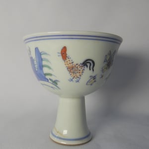 May include: White porcelain goblet with a pedestal base, adorned with hand-painted illustrations. The design includes a rooster, blue foliage, and floral motifs. The rim and base are accented with blue stripes, creating a classic aesthetic.