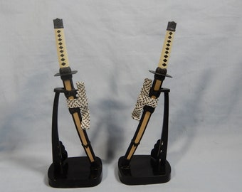 Könnte beinhalten: Zwei Miniatur-Katana-Schwerter mit schwarzen Griffen und Ständern. Die Klingen sind beige mit schwarzen Akzenten, und die Griffe sind mit cremefarbenem Material umwickelt. Jedes Schwert wird auf einem schwarzen Ständer präsentiert.