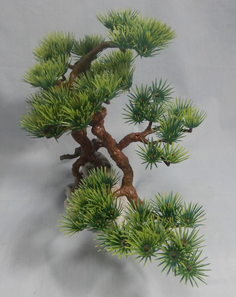 Op de afbeelding: Kunstmatige bonsai boom met groene naalden en bruine takken. De boom is bevestigd op een witte rotsbasis. De takken zijn kunstzinnig gerangschikt, waardoor de indruk van een miniatuurboom ontstaat. Het ontwerp is bedoeld voor decoratieve doeleinden.