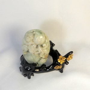 May include: A carved jade egg with intricate details, resting on a black wooden stand with gold accents. The stand is shaped like a tree branch with stylised flowers.