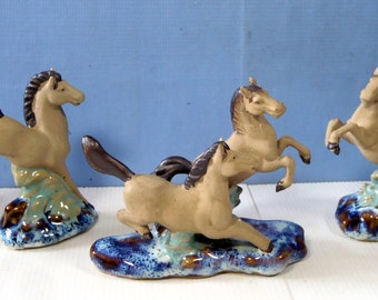 May include: Four ceramic horse figurines, each standing on a blue and brown base. The horses are all in a leaping pose, with their manes and tails flowing in the wind.