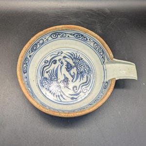 May include: A round, ceramic ashtray with a blue and white phoenix design in the center. The ashtray has a light brown rim and a rectangular rest. The design is reminiscent of traditional Asian art.