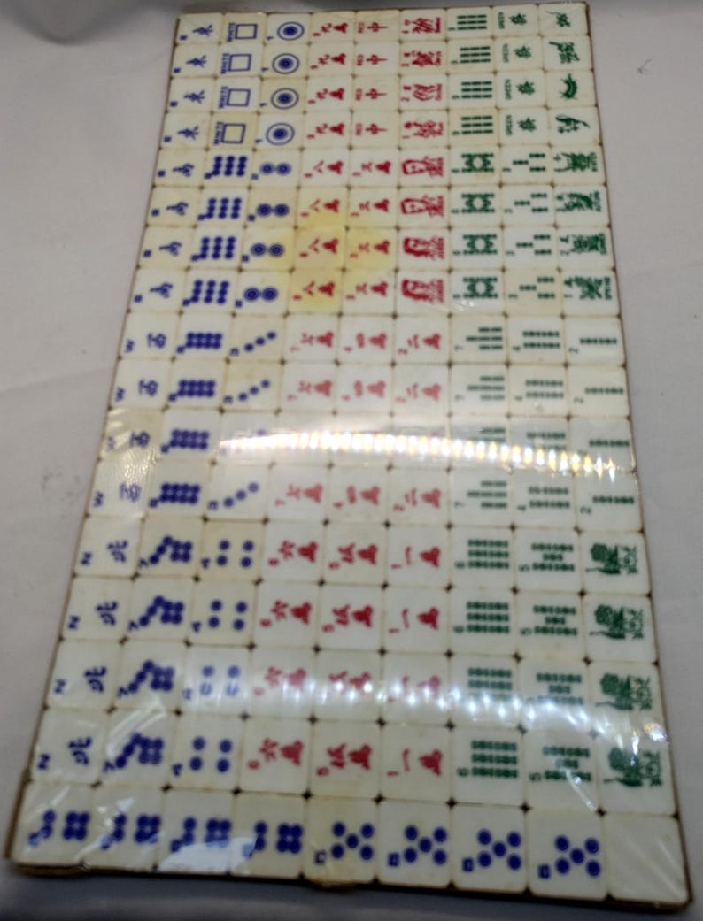 May include: A complete set of vintage Mahjong tiles, arranged in a rectangular pattern. The tiles are off-white with symbols in red, green, and blue. The set is packaged in clear plastic, suggesting it is a complete game set.