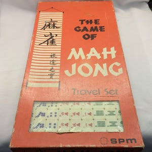 May include: An orange Mah Jong travel set box with the game tiles visible through a clear window. The box has text in English and Chinese characters. The tiles are white with red and blue markings. The box is rectangular.