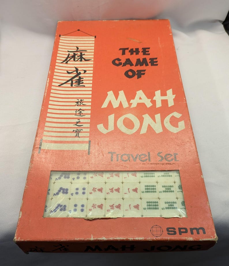 May include: An orange Mah Jong travel set box with the game tiles visible through a clear window. The box has text in English and Chinese characters. The tiles are white with red and blue markings. The box is rectangular.