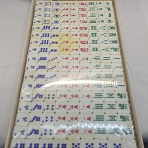 May include: A vintage Mahjong set in a cardboard box. The tiles are white with various symbols in red, green, blue, and black. The set appears to be complete and ready for play. The box is beige.