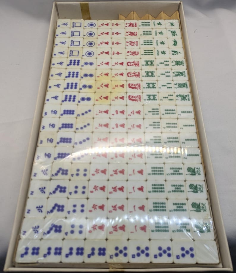 May include: A vintage Mahjong set in a cardboard box. The tiles are white with various symbols in red, green, blue, and black. The set appears to be complete and ready for play. The box is beige.