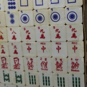 May include: A complete set of Mahjong tiles, featuring white tiles with various symbols and characters in red and blue. The set includes tiles with numbers, suits, and special characters, all neatly arranged in a box.