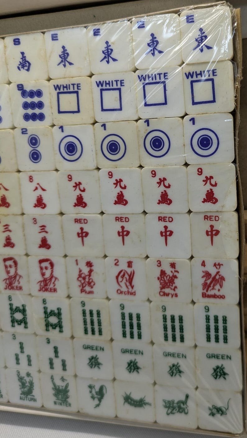 May include: A complete set of Mahjong tiles, featuring white tiles with various symbols and characters in red and blue. The set includes tiles with numbers, suits, and special characters, all neatly arranged in a box.