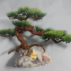 Op de afbeelding: Een miniatuur bonsai boomsculptuur met een bruine stam en takken, groen blad en een klein figuur in gele kleding zittend op een rotsformatie. De sculptuur is een decoratief item.