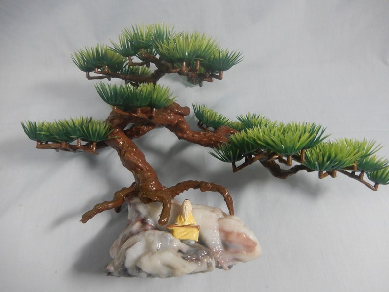 Op de afbeelding: Een miniatuur bonsai boomsculptuur met een bruine stam en takken, groen blad en een klein figuur in gele kleding zittend op een rotsformatie. De sculptuur is een decoratief item.