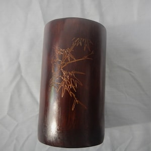 Puede incluir: Un jarrón de bambú marrón oscuro con un diseño tallado de ramas de bambú. El jarrón mide aproximadamente 15 cm de alto.