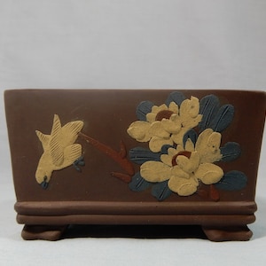 May include: A rectangular brown ceramic planter with a raised base. The planter features a painted design of a yellow bird and white flowers with blue leaves.