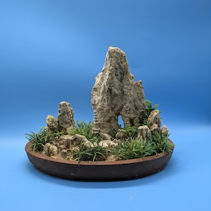 May include: A miniature bonsai landscape featuring a rock formation with an arch, surrounded by small green plants and sand. The arrangement is set in a brown, oval-shaped pot, creating a serene and natural aesthetic.
