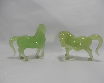 May include: Two jade green horse figurines. The horses are standing on a white surface.