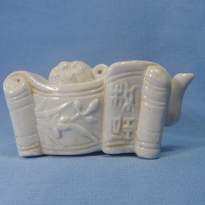 Puede incluir: Tetera de cerámica blanca con forma de rollo y un pequeño pico. La tetera tiene un diseño floral y caracteres chinos en el lateral.