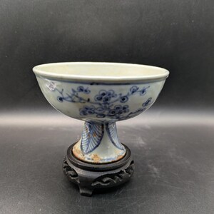 Puede incluir: Un cuenco de cerámica blanco y azul con un diseño floral, montado sobre una base de pedestal. El cuenco está sostenido por un soporte decorativo de madera negra. El diseño del cuenco presenta patrones florales azules.