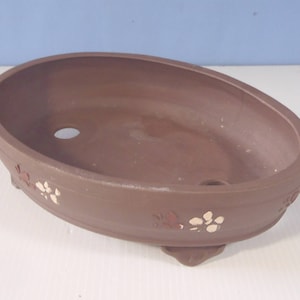 May include: A brown oval ceramic bonsai pot with white and brown floral designs. The pot has three small feet.