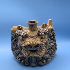 May include: A ceramic vase in the shape of a lion's head, with a tan and gray glaze. The vase has a cylindrical opening at the top and is set against a blue background. The lion's face is detailed with eyes, a nose, and a mouth.
