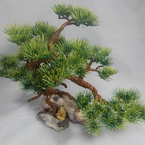 Op de afbeelding: Kunstmatige bonsai boom met groene naalden en bruine takken. De boom is gemonteerd op een rotsformatie met een klein geel beeldje. De bonsai is een decoratief item.