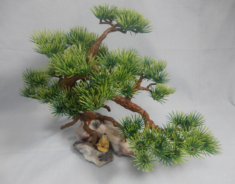 Op de afbeelding: Kunstmatige bonsai boom met groene naalden en bruine takken. De boom is gemonteerd op een rotsformatie met een klein geel beeldje. De bonsai is een decoratief item.